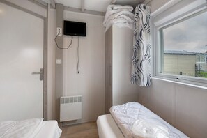 Een kleine, eenvoudige accommodatie met een bed, een kachel, een aan de muur bevestigde televisie en een raam met uitzicht op een wateroppervlak.