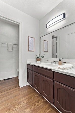 A bathroom with a wooden floor, white walls, a large mirror, a sink with a soap dispenser, and a towel rack. - The Grove on 66th- 3B 2Bth Tulsa Duplex (Tulsa)