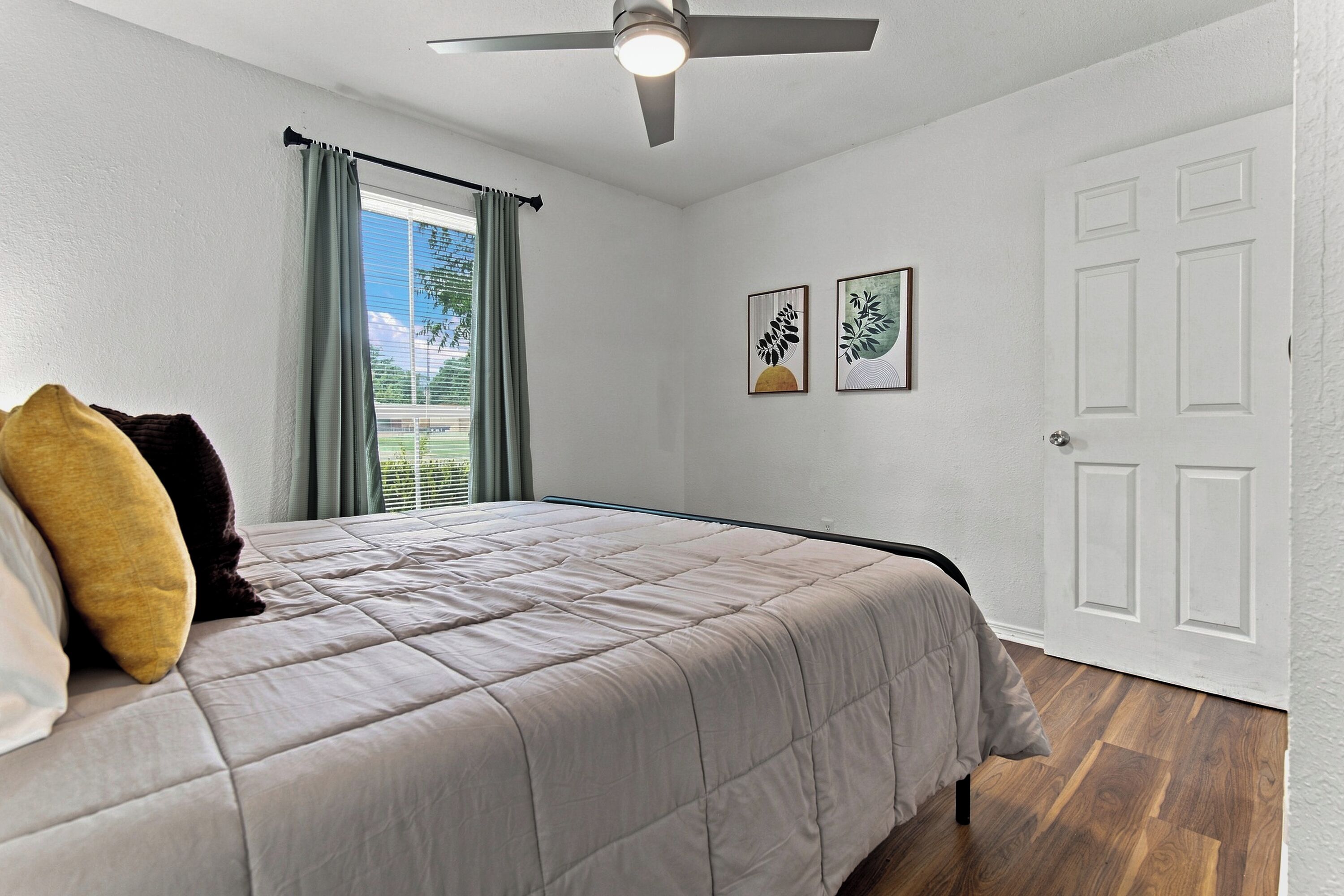 Une chambre à coucher avec un lit, un ventilateur de plafond, une fenêtre avec des rideaux et deux tableaux encadrés accrochés au mur.