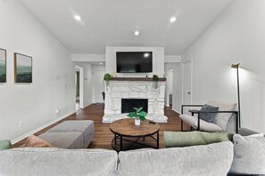 A modern living room with a stone fireplace, a flat-screen TV mounted above it, a round coffee table with a plant, and a grey sofa with cushions.