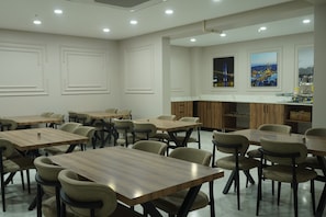 A modern dining area with multiple tables and chairs, a kitchen area with wooden cabinets, and a cityscape view through the windows. - ÇELİK PALACE HOTEL (İstanbul)