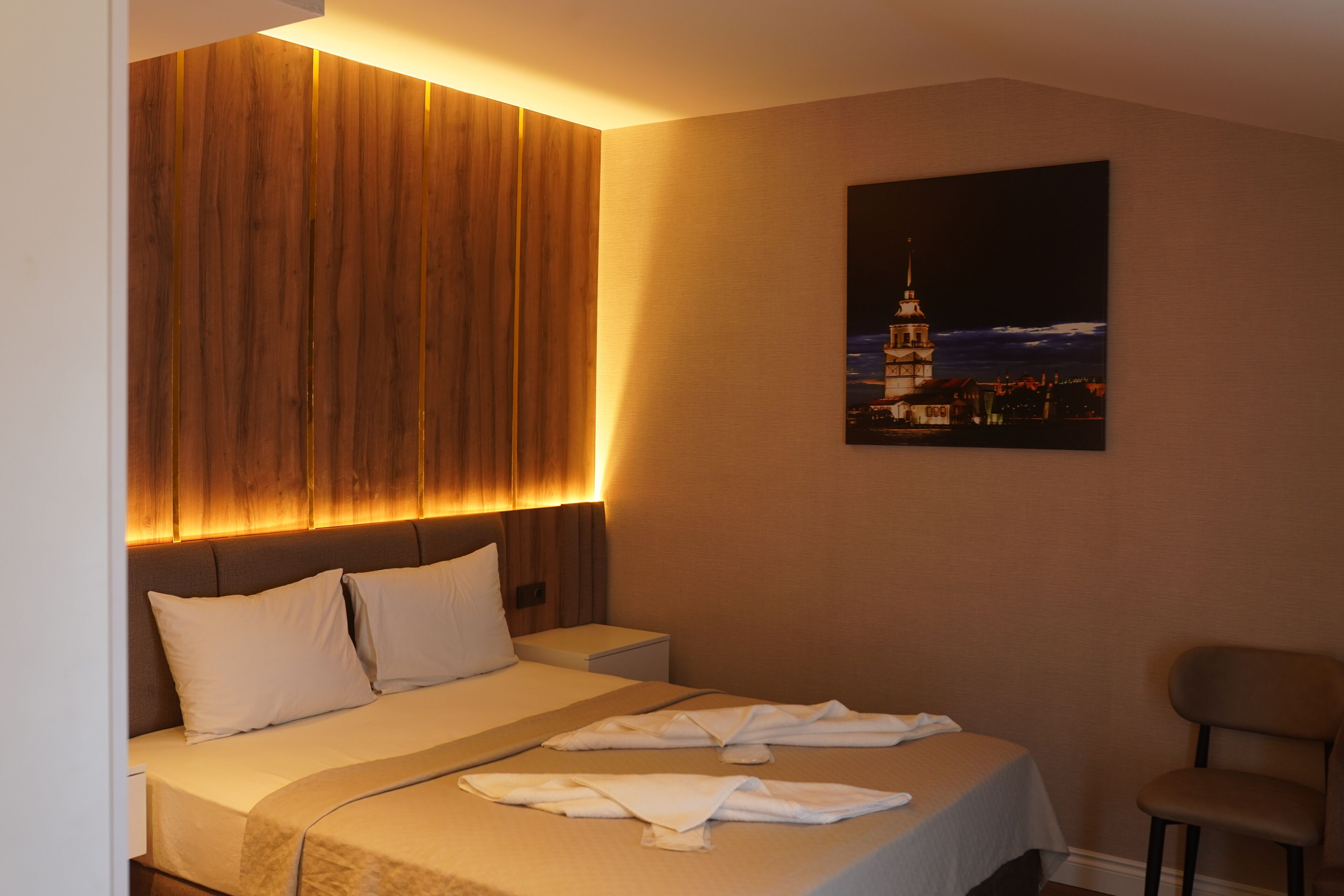 A hotel room with a large bed, a wooden headboard, a nightstand, a chair, and a painting of a cityscape.