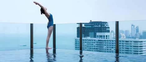 Una persona con traje de baño azul realizando una postura de yoga en una piscina infinita con fondo de cristal y un horizonte urbano al fondo.