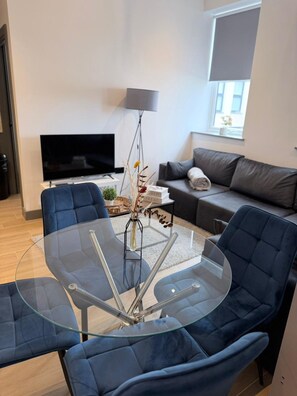 A modern living room with a glass-top coffee table surrounded by blue chairs, a grey sofa, a floor lamp, and a flat-screen TV. - Modern 2BR Flat | North Finchley (Greater London)