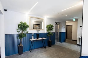 A reception area with a blue and white color scheme, a reception desk, two potted plants, a mirror, and an elevator. - Modern 2BR Flat | North Finchley (Greater London)