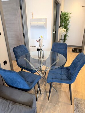 A modern dining area with a glass table surrounded by blue upholstered chairs, a white wall-mounted air conditioning unit, and a potted plant. - Modern 2BR Flat | North Finchley (Greater London)