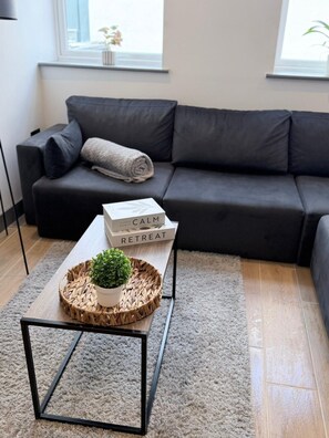 A modern living room with a dark grey sofa, a small coffee table with a plant and a book titled 'RETREAT,' and a light grey rug. - Modern 2BR Flat | North Finchley (Greater London)