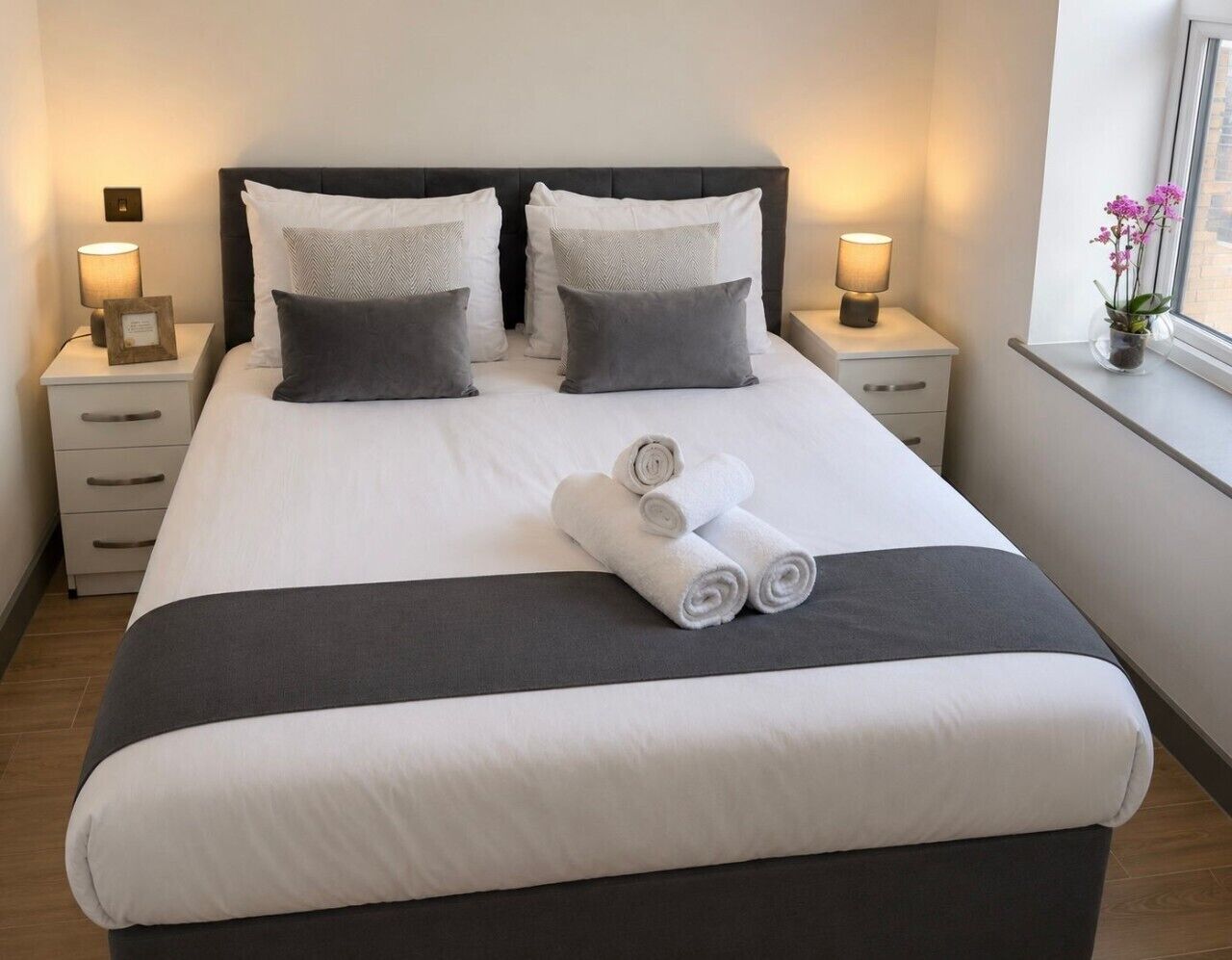 A neatly made bed with white and gray linens, two bedside tables with lamps, and a window with a view.