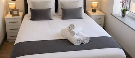 Una cama bien hecha con ropa de cama blanca y gris, dos mesitas de noche con lámparas y una ventana con vistas.