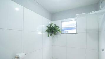Casa de banho com azulejos brancos, janela, planta e porta-papel higiénico.