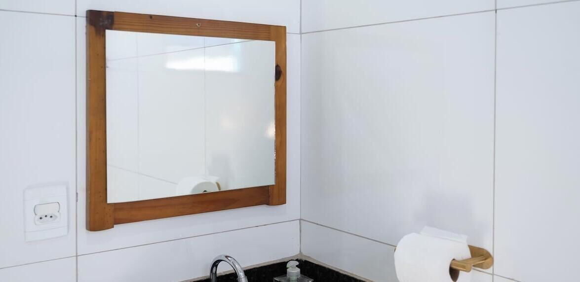 Ein Badezimmer mit weißen Fliesen, einem spiegel mit Holzrahmen, einem Waschbecken mit Armatur und einem Toilettenpapierhalter.