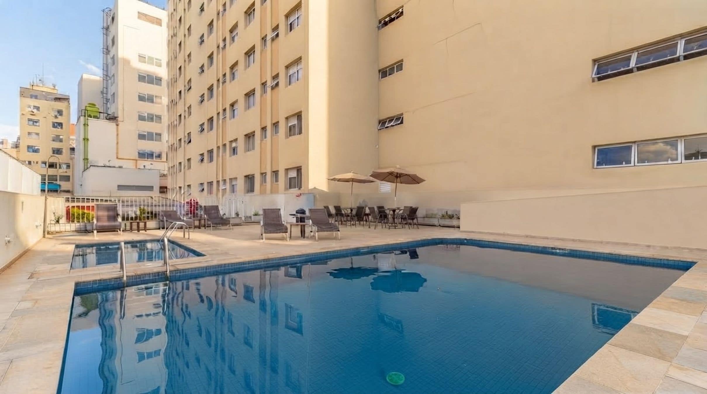 A swimming pool with lounge chairs and umbrellas, surrounded by a high-rise building.