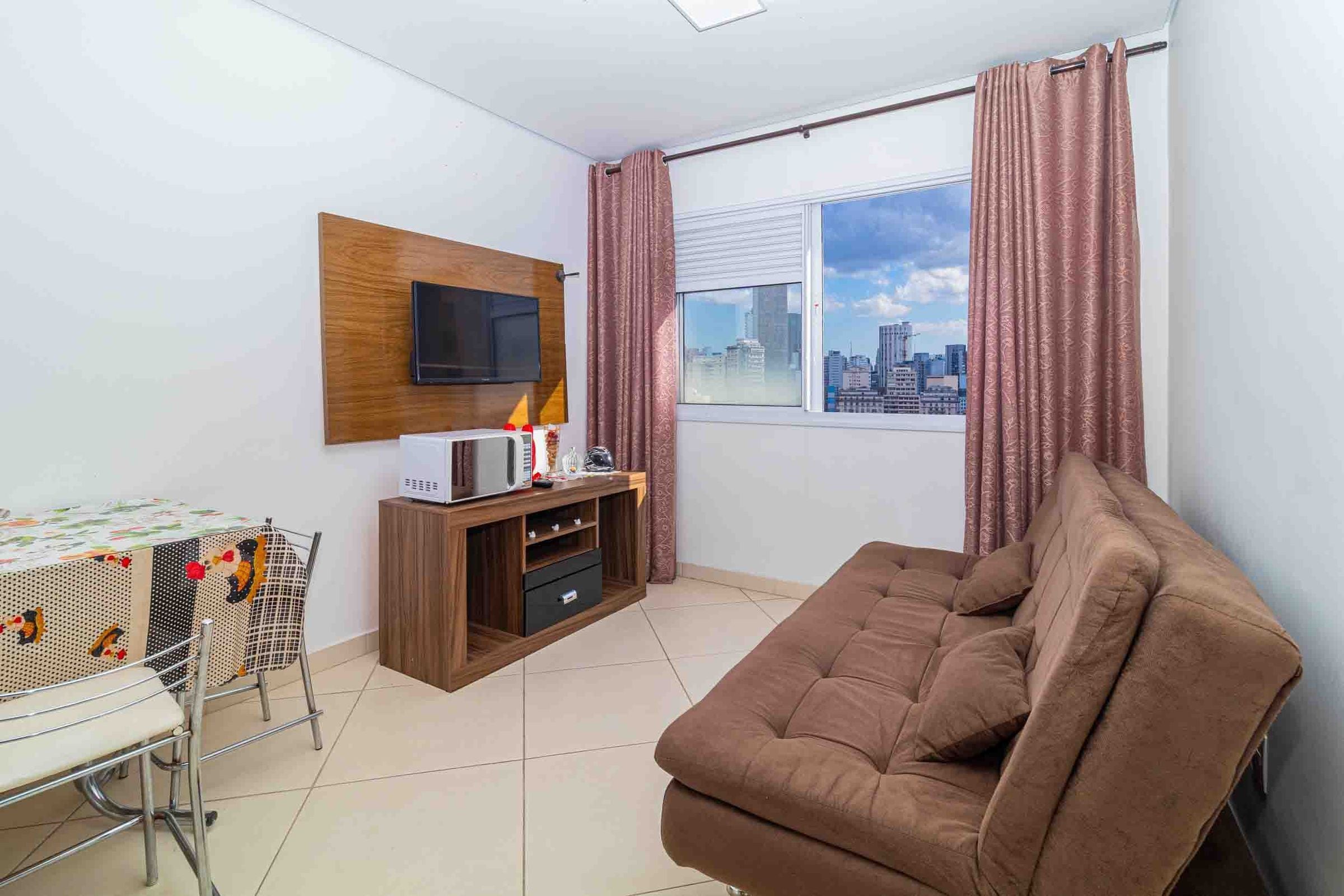 Een woonkamer met een bruine bank, een tv bevestigd aan een houten paneel, een klein tafeltje met een tafelkleed met patroon, en een raam met gordijnen.