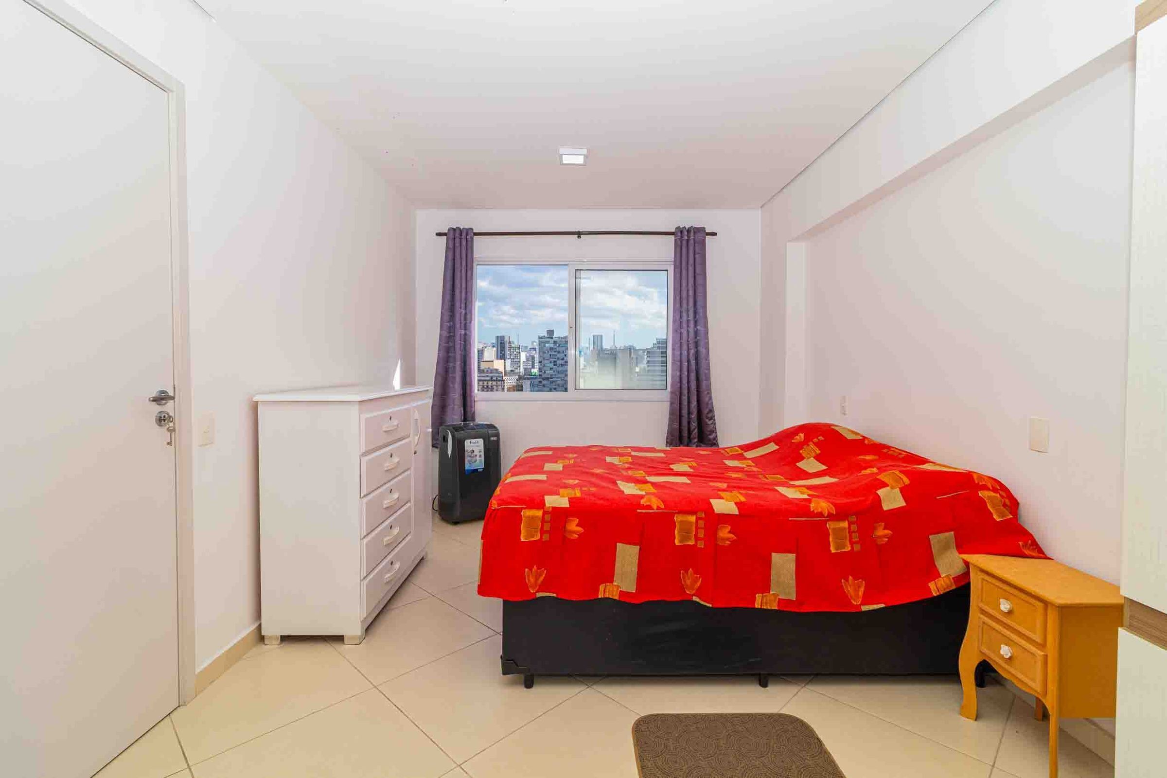 Een slaapkamer met een bed, een dressoir, een koffer en een raam met gordijnen.