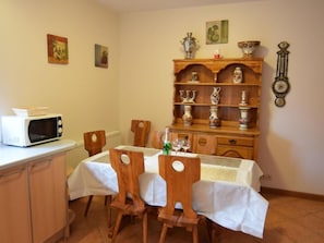 Un coin repas avec une table dressée pour quatre personnes, des chaises en bois, une nappe blanche, un meuble contenant un four à micro-ondes et une étagère avec des objets décoratifs.
