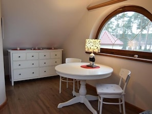 A room with a white round table, a white chair, a white cabinet with drawers, and a window with a view of trees.