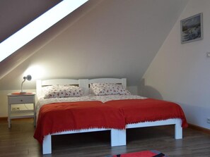 Quarto com teto inclinado, cama com manta vermelha, mesa de cabeceira com abajur e um quadro na parede.