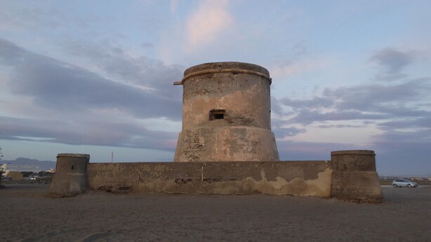 Una torre cilíndrica con una pequeña ventana, rodeada por un muro bajo, frente a un cielo nublado.