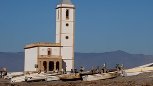 Una iglesia con un campanario y un reloj, rodeada de barcos en una orilla arenosa.