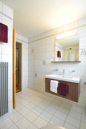 Ein Badezimmer mit weiß gefliestem Boden, einem Holzwaschtisch und einem Spiegel sowie zwei Handtüchern, die an der Wand hängen.