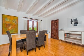 A dining area with a wooden table and wicker chairs, a painting on the wall, a window with curtains, and a bookshelf with books. - 6-guest Stone Haven in Pollensa (Pollença)