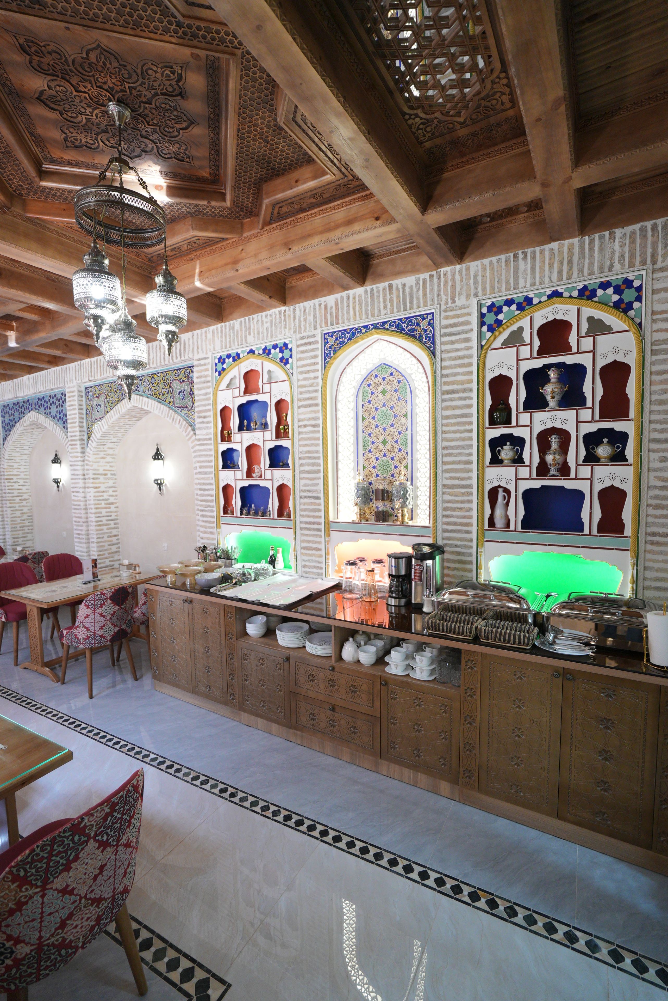 Un interior tradicional con techo de madera, decoraciones murales ornamentales, una zona de bar con copas y un área de estar con cojines estampados.