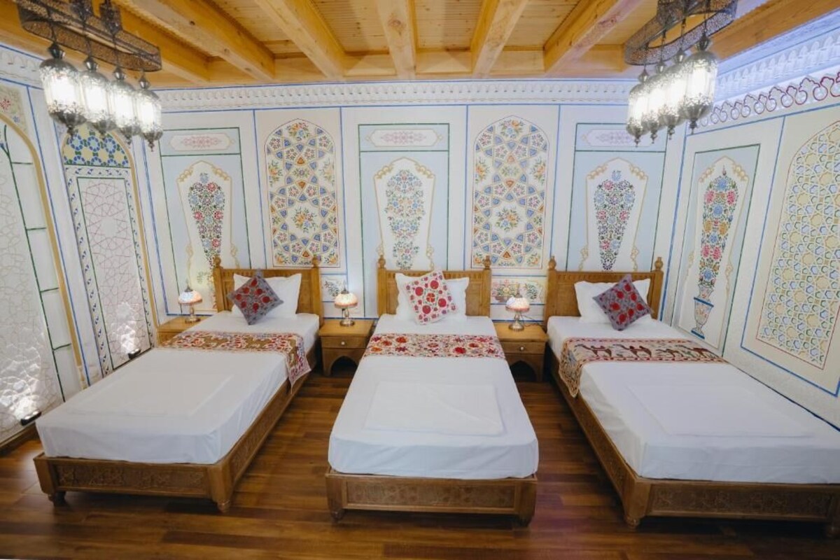 A room with three wooden beds, each with a white bedspread and decorative pillows. The room has ornate wall decorations and a wooden ceiling.