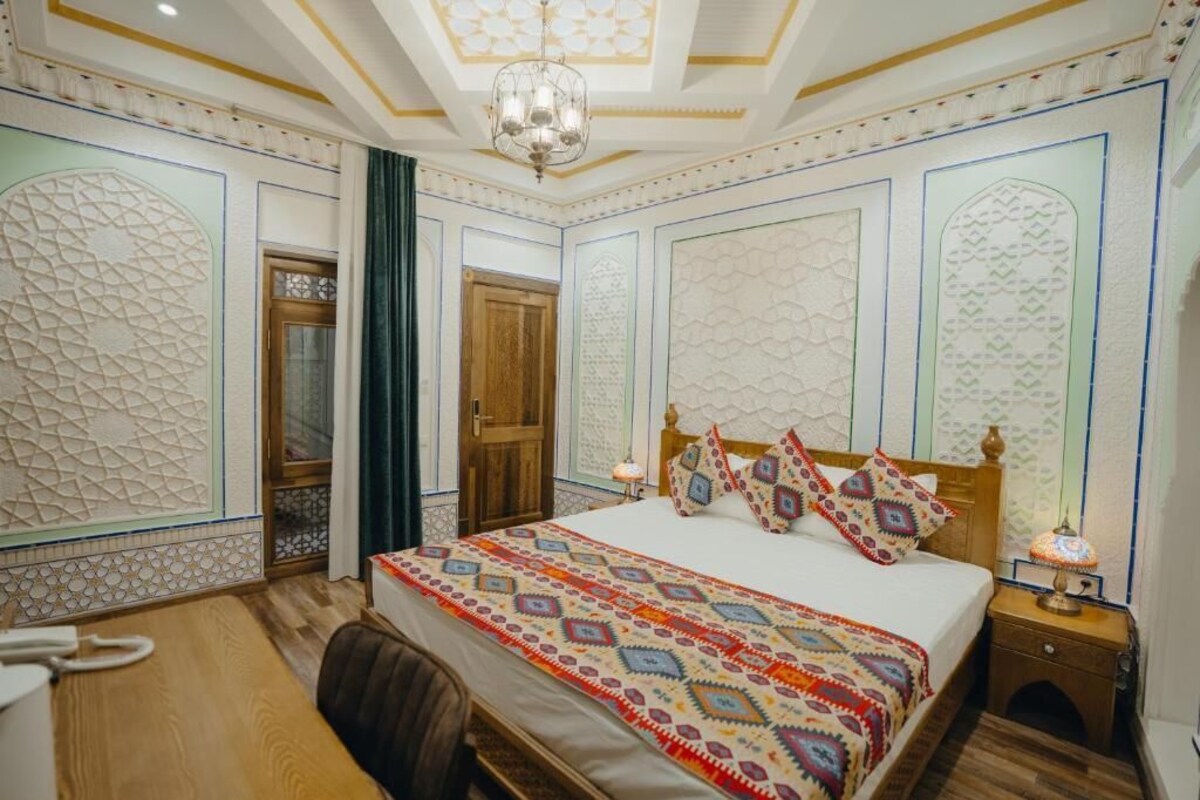 A bedroom with a large bed, a wooden headboard, patterned wall decorations, a chandelier, and a nightstand with a lamp.