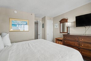 A hotel room with a bed, a dresser, a television, a painting, and a door. - Ocean View 1BR Suite + Beach Access + Lazy River (Myrtle Beach)