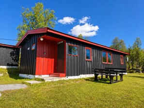 Ein kleines, eingeschossiges Gebäude mit roter Tür und Fenstern, umgeben von Gras und Bäumen.