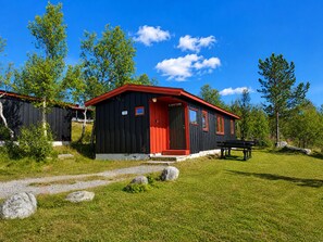 Une cabane noire et rouge, avec une porte rouge, portant le numéro 190, entourée d’herbe et d’arbres.