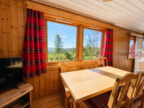 Un interior de madera con una mesa de comedor, sillas, un televisor y ventanas con cortinas rojas.