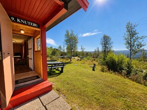 Entrada a una cabaña con fachada de madera, un banco y vistas a un lago y árboles.