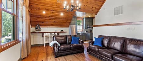 A spacious living room with a high ceiling and wooden beams, furnished with leather sofas and a chandelier.