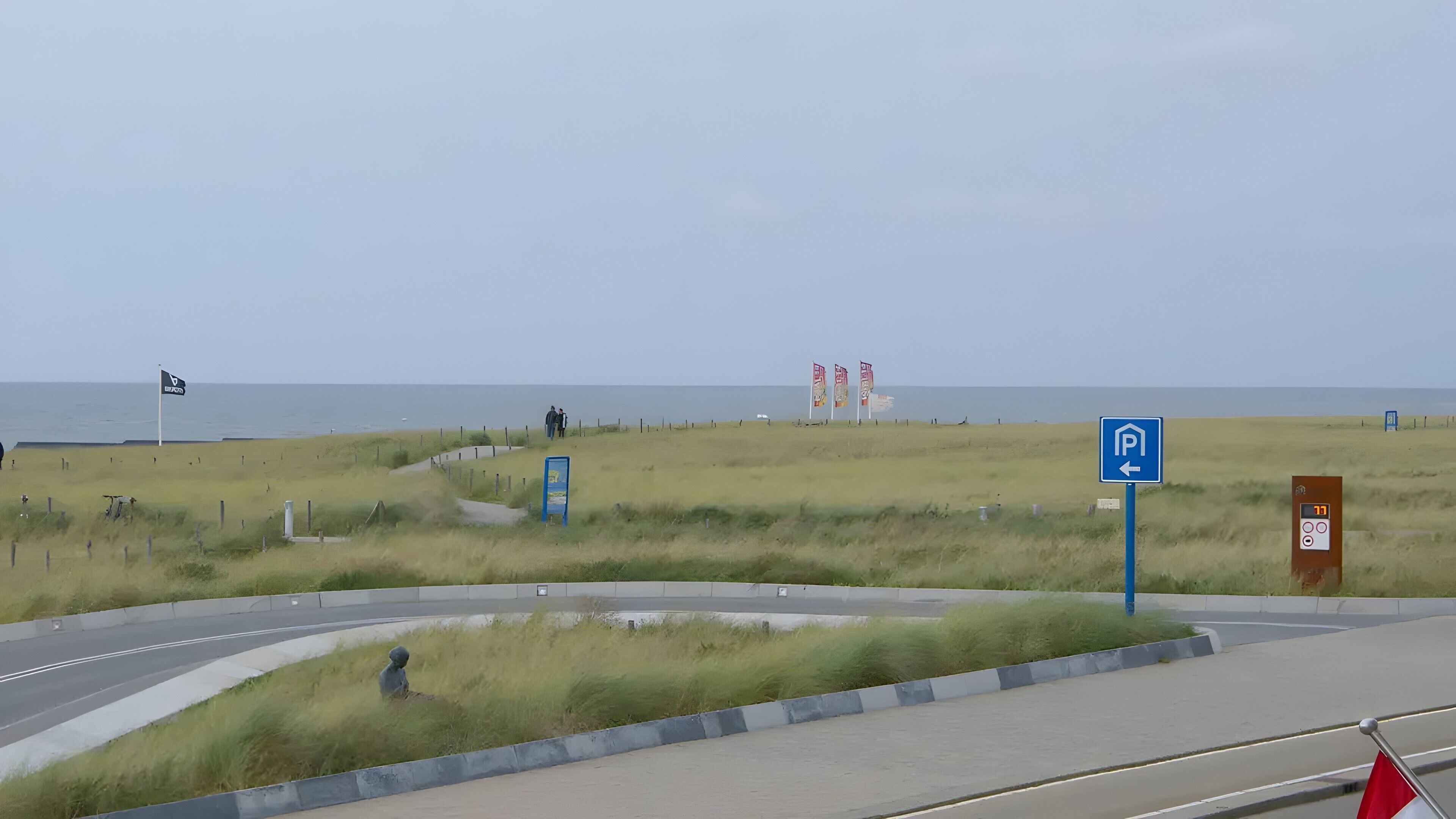 Eine Küstenstraße mit Parkschild, Fußweg und Strand im Hintergrund.