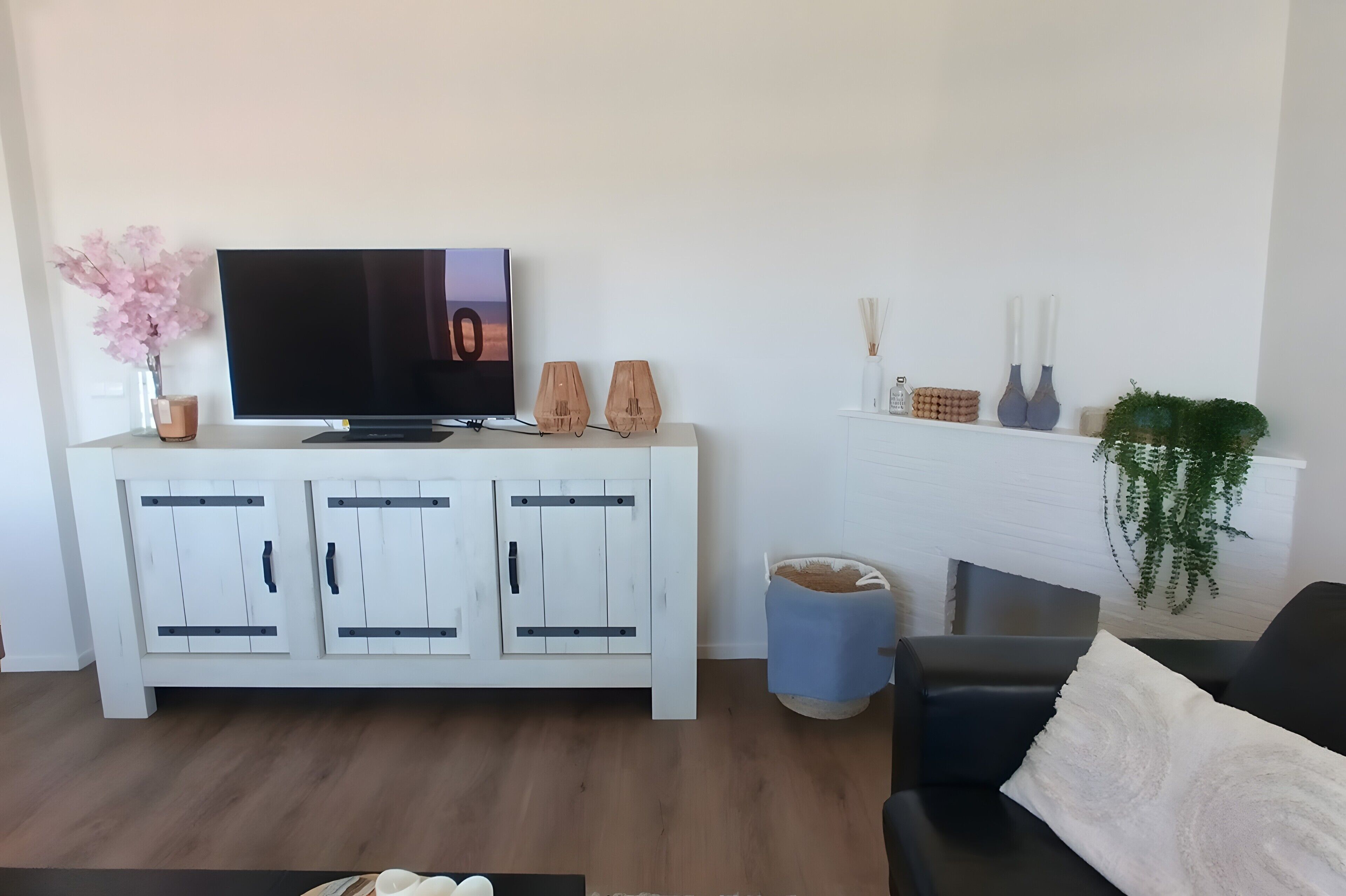 Ein Wohnzimmer mit Flachbildfernseher, weißen Schränken, einem blauen Hocker, einer schwarzen Couch und einem weißen Kaminvorsatz mit Dekoration.
