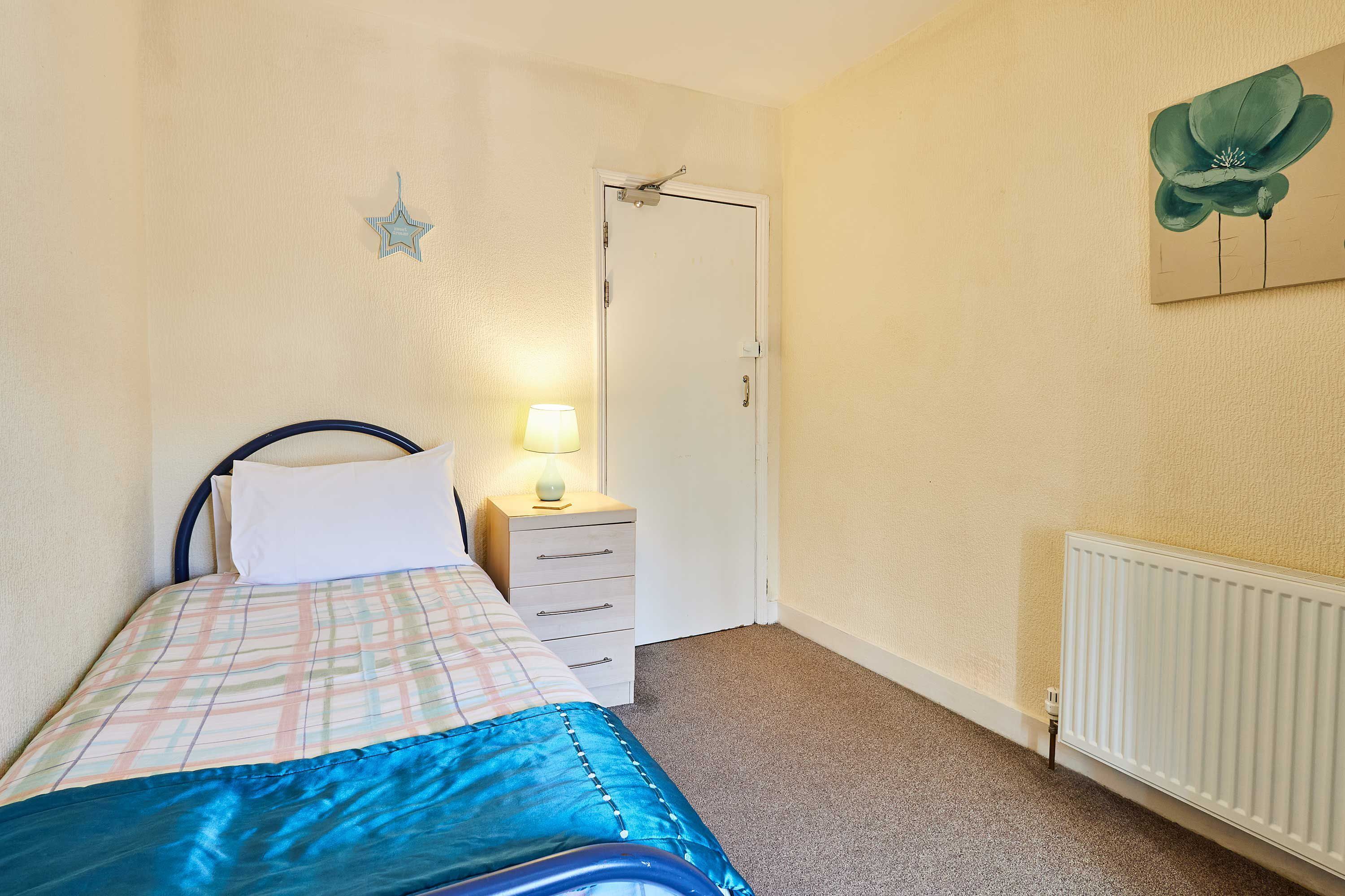 Een slaapkamer met een bed, nachtkastje, lamp en een deur.