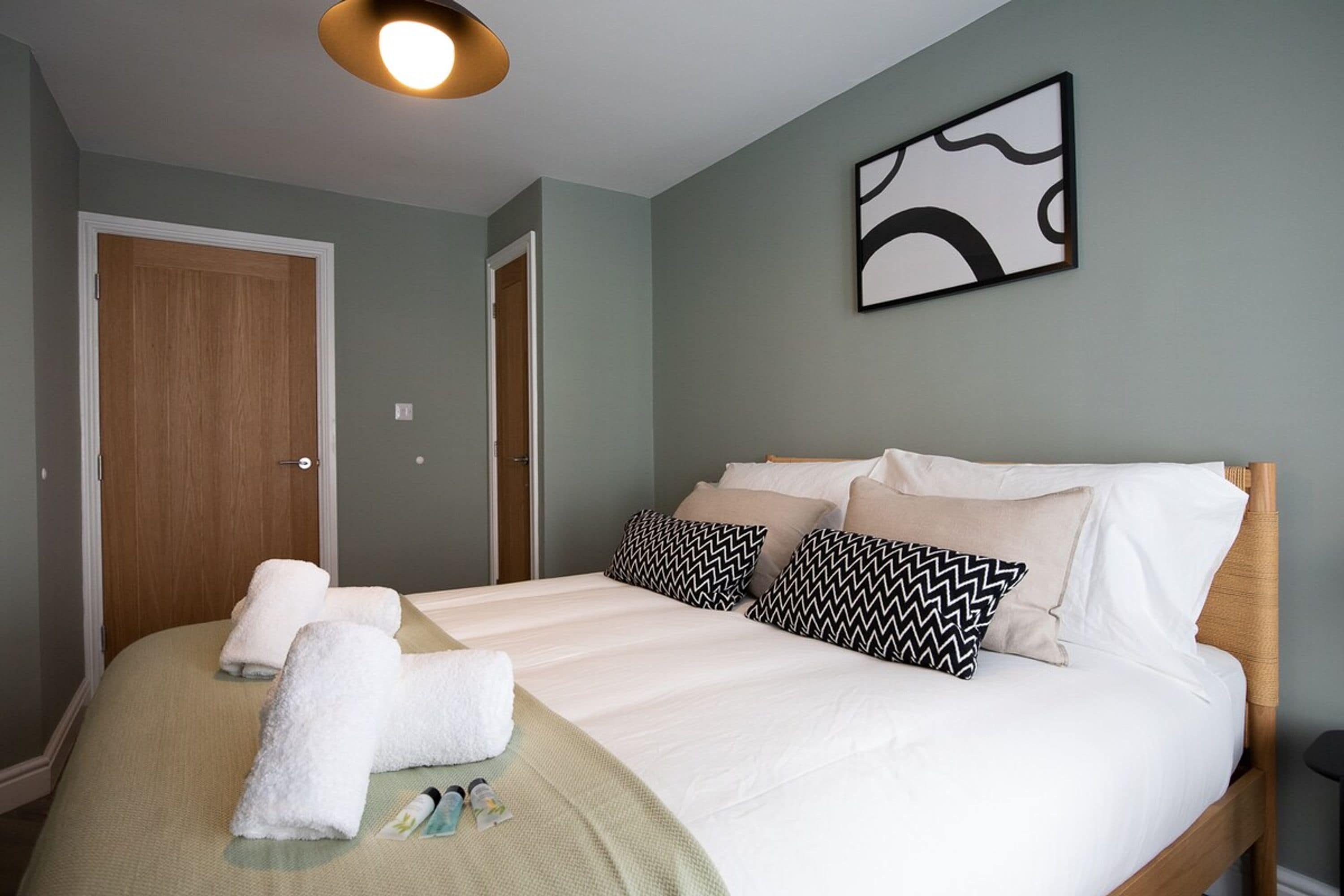 A neatly made bed with white linens and two patterned pillows, a wooden door, and a wall-mounted abstract art piece.
