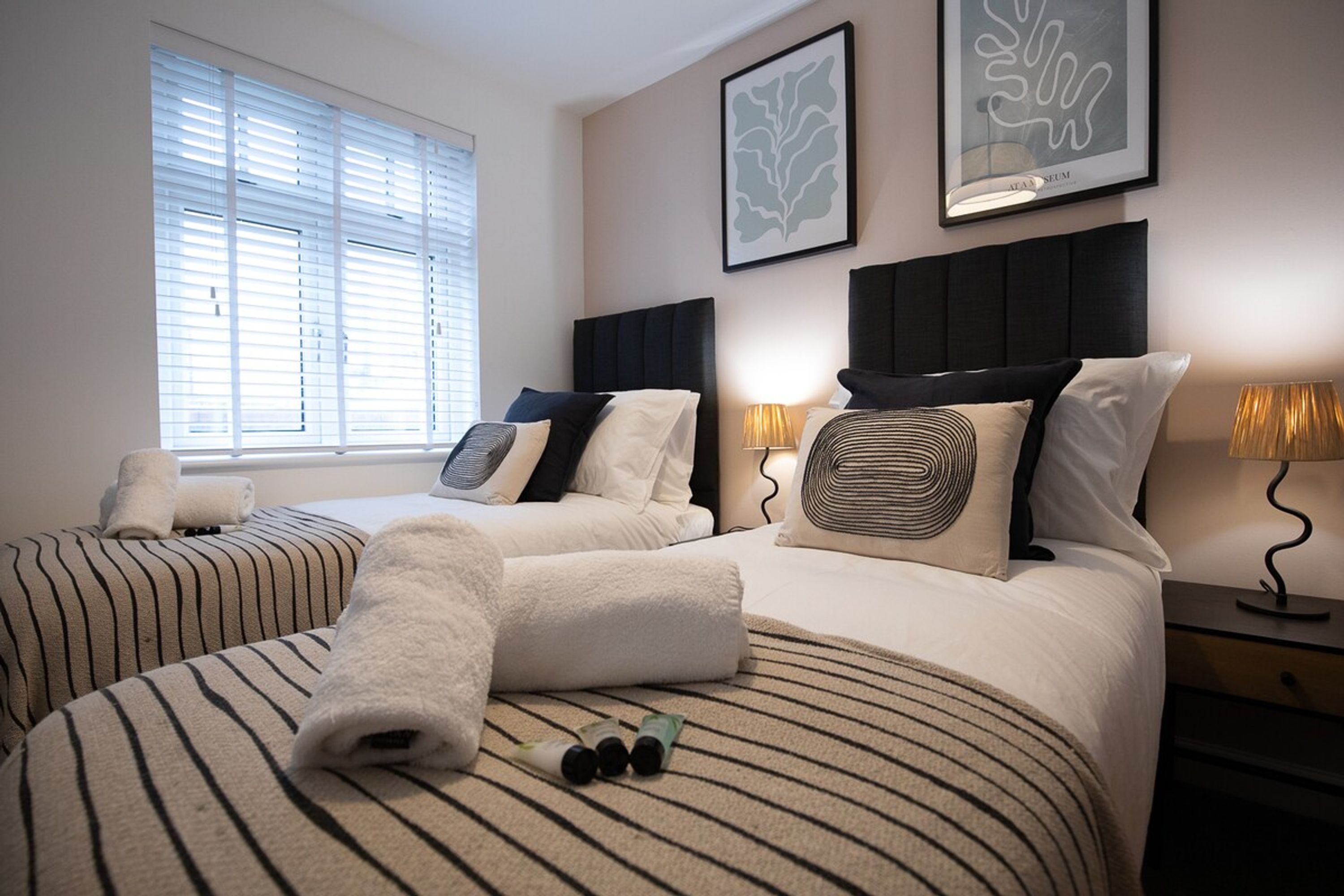 Een hotelkamer met twee bedden, een raam met zonwering, een nachtkastje met een lamp en kunst aan de muur.