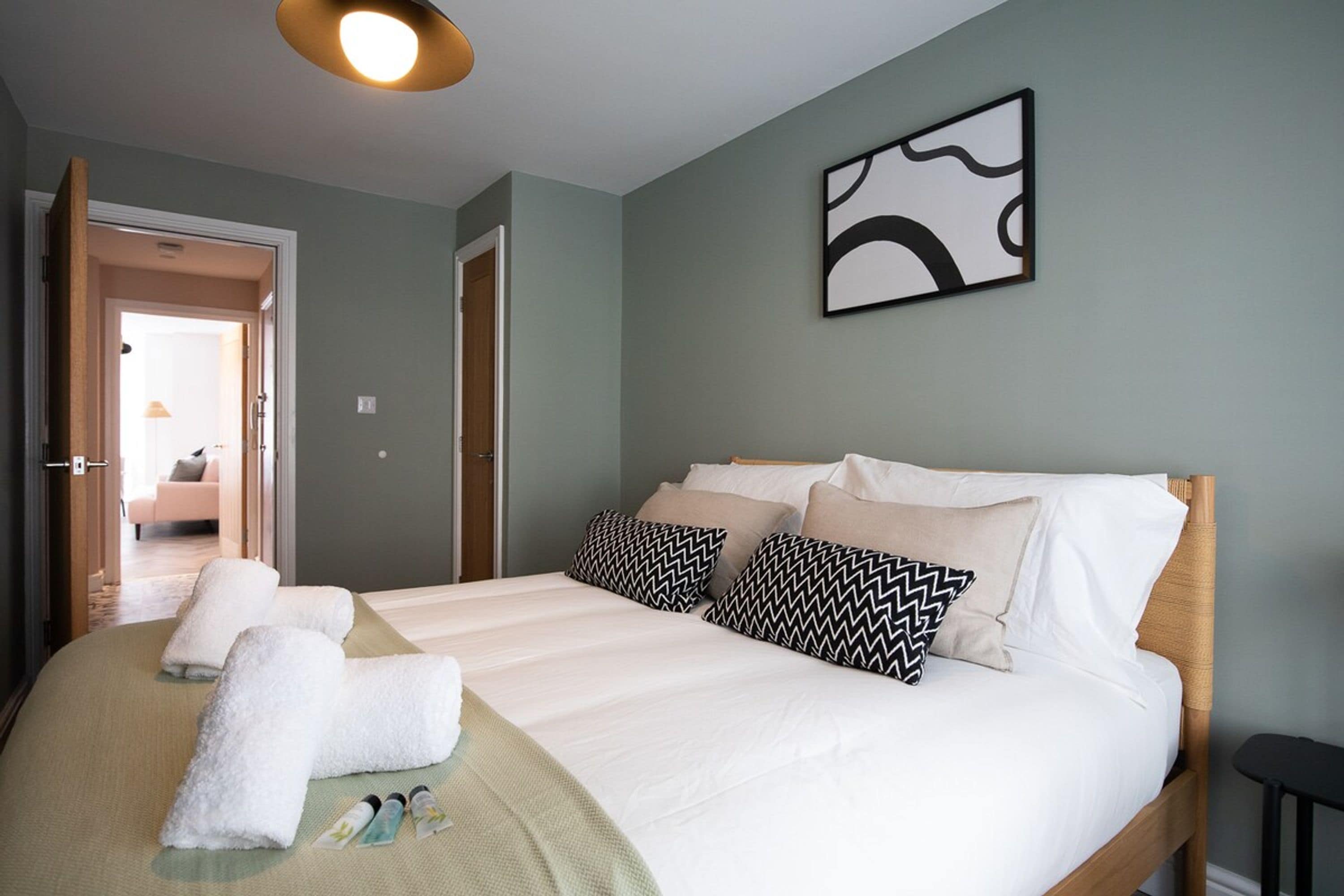 Una cama bien tendida con sábanas blancas y almohadas estampadas, una mesita de noche y una obra de arte abstracto colgada en la pared.