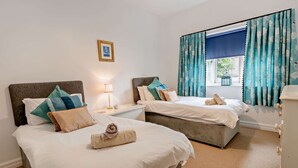 Quarto de hotel com duas camas, uma mesa de cabeceira com luminária, um quadro na parede e uma janela com cortinas.