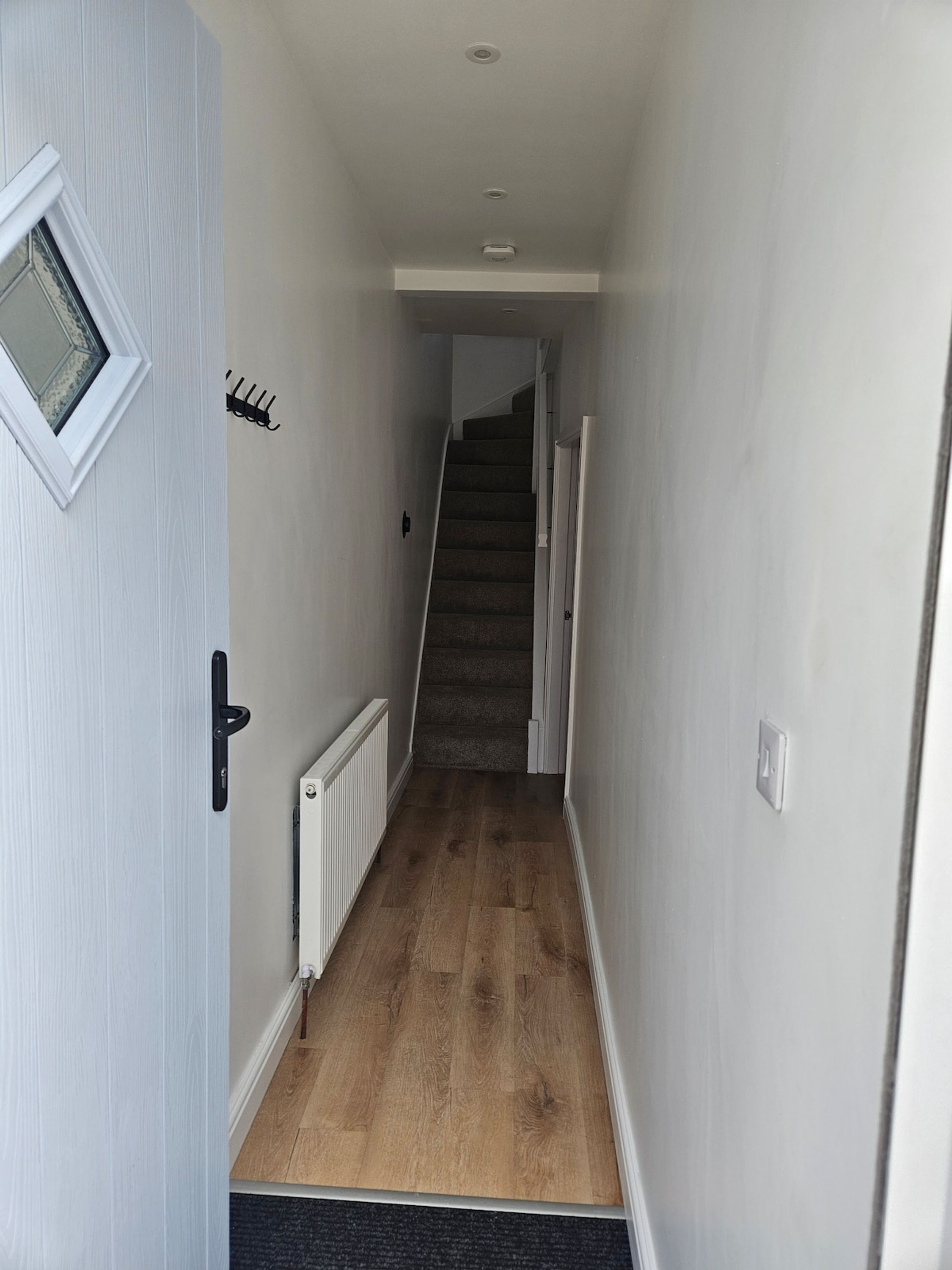 A narrow hallway with a wooden floor, a door with a window, and a staircase in the distance.