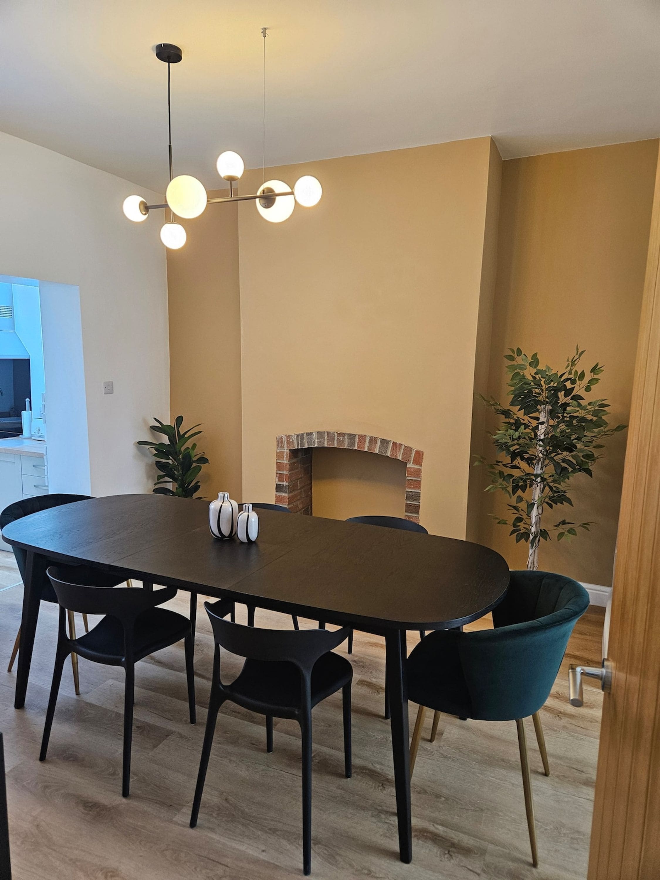 A dining area with a round table, black chairs, a modern chandelier, a brick fireplace, and a potted plant.