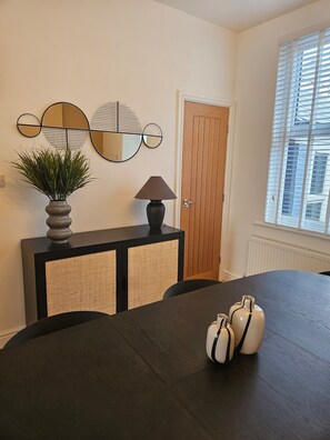 A dining area with a table, chairs, a console table with a lamp, a potted plant, and a wall-mounted mirror. - Salt Sage - Whitby (Whitby)