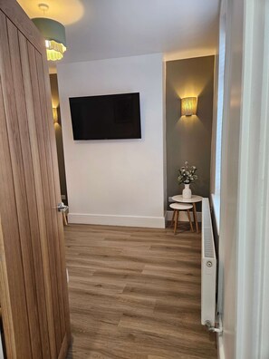 Ein Flur mit einem an der Wand befestigten Flachbildfernseher, einem kleinen runden Tisch mit einer Vase und einem Heizkörper auf dem Boden.