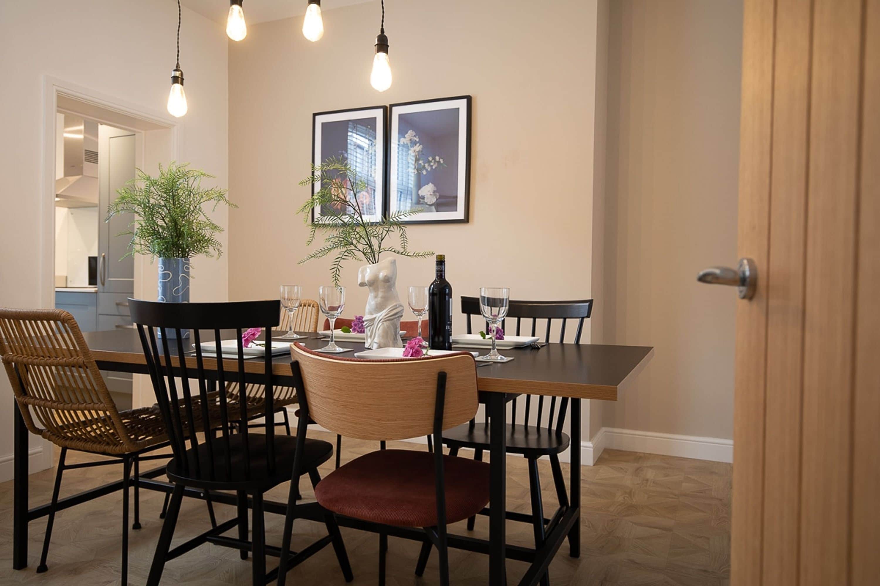A dining area with a table set for two, chairs, a potted plant, and framed pictures on the wall.