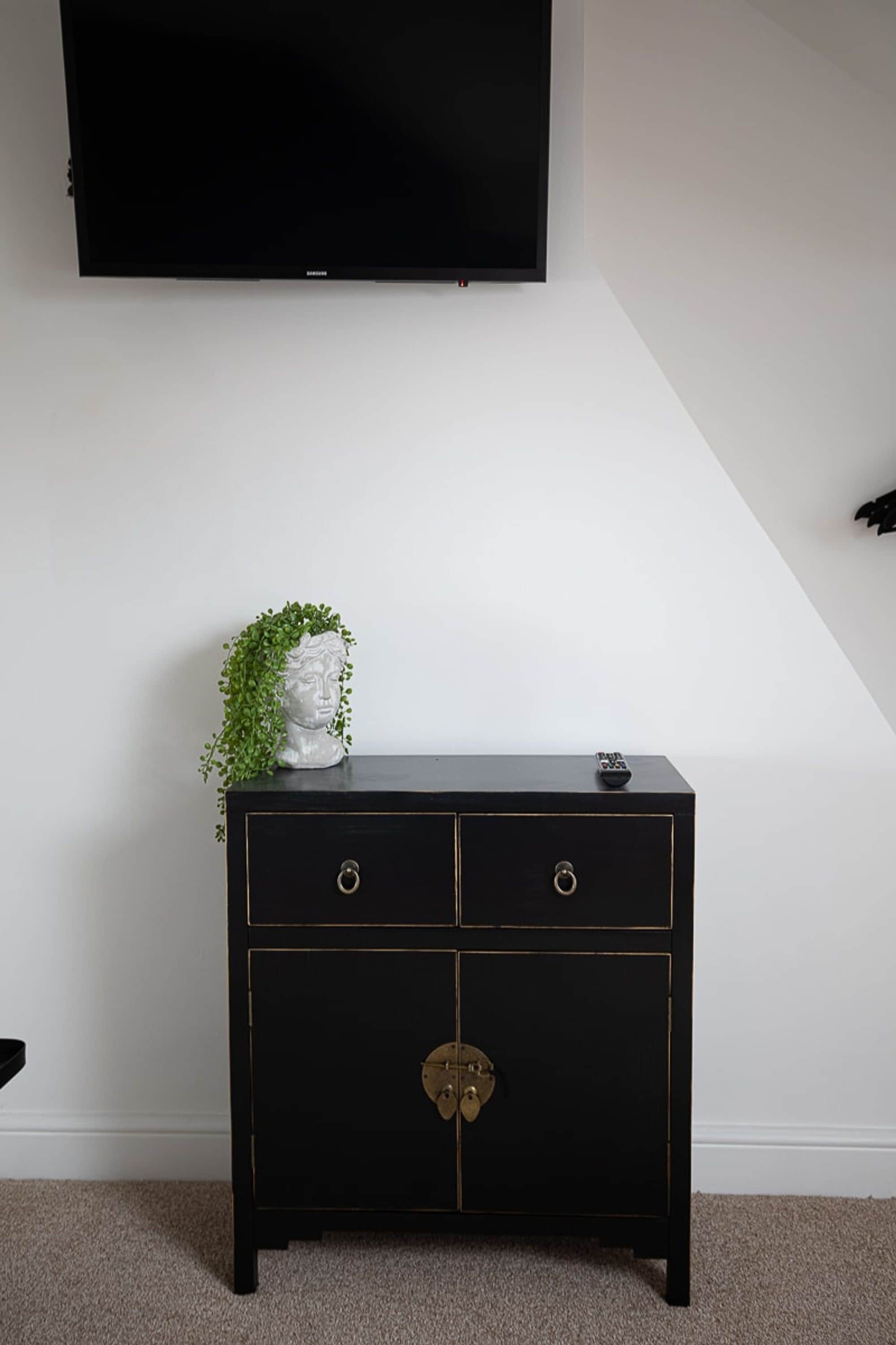Eine schwarze Kommode mit zwei Schubladen und einer abschließbaren Schranktür, an einer weißen Wand platziert, auf der ein Flachbildfernseher angebracht ist.