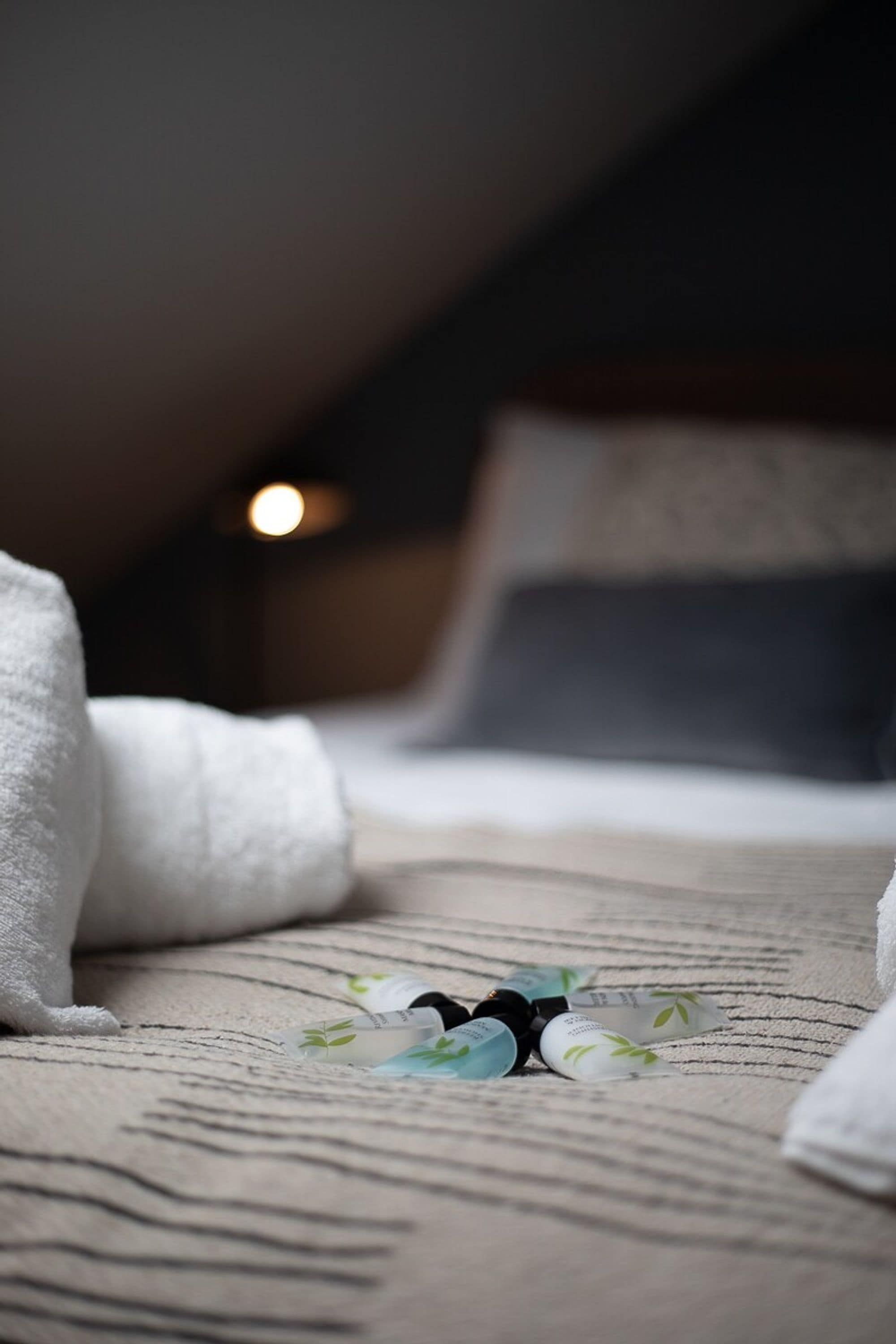 Een opgemaakt bed met een gestreept dekbed en kussens, een opgerolde witte handdoek en een flesje lotion op het bed.