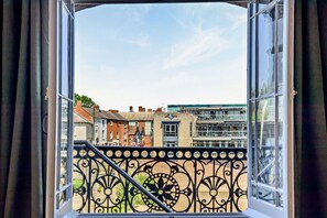Blick aus einem Fenster mit einem schmiedeeisernen Balkongeländer auf eine Stadtansicht mit Gebäuden und blauem Himmel.