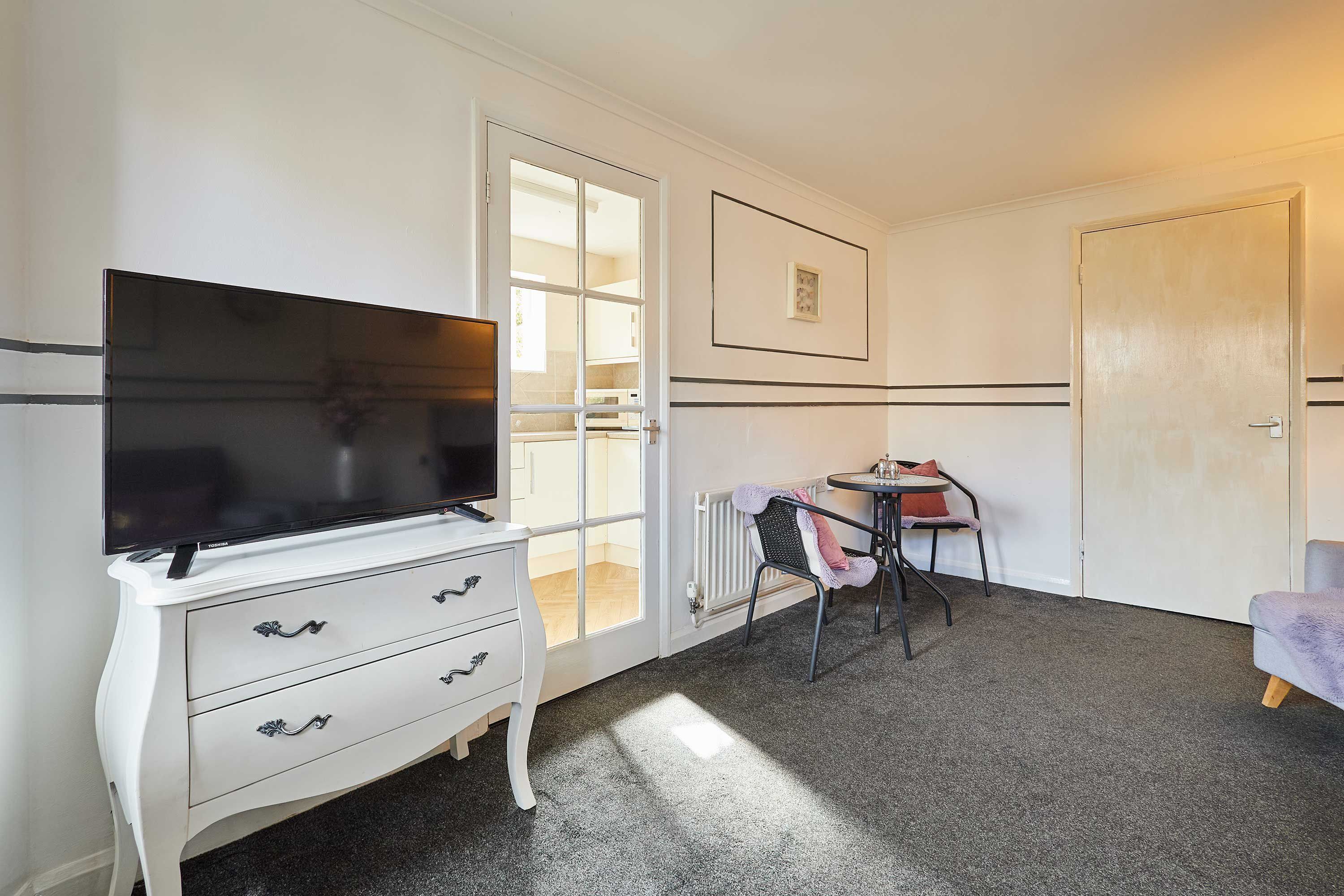 A room with a white dresser, a flat-screen TV, a small round table with chairs, and a door.